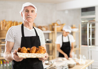 Experienced elderly baker in uniform stands in small private bakery offering croissants