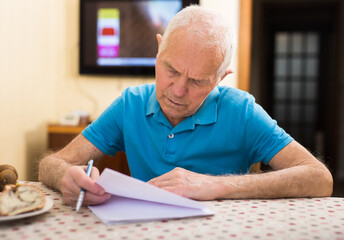 Elderly man writes a letter on a sheet of paper while sitting at a table in room