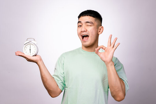 A cheerful Asian man in a mint green shirt holds a white alarm clock in one hand and makes an "OK" gesture with the other, smiling confidently against a white background