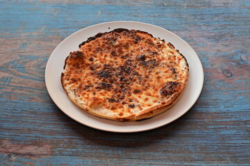 A pizza with cheese and toppings sits on a white plate on a wooden table