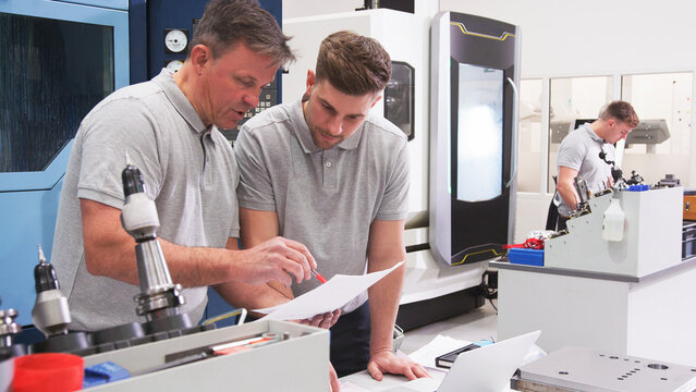 Male Engineer And Apprentice Discussing Plans For Job Whilst Looking At Laptop