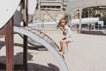 A girl climbs a blue rope net on a weathered wooden playground by the beach. Barefoot and determined, she enjoys a sunny summer day full of adventure and fun.
