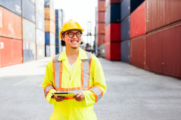 Smiling logistics Asian worker in high-visibility safety suit and hard hat holds a tablet while inspecting shipping containers at a cargo port. Global trade, freight management, supply chain logistics