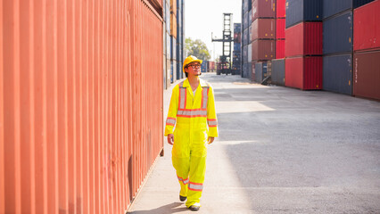 Asian logistics worker in high-visibility safety suit and hard hat walks through a busy shipping...