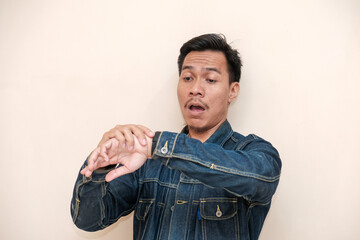 Obraz premium A Southeast Asian man wearing a denim jacket shocked expression to see watch on hand against a white background. This image is perfect for content related to time management
