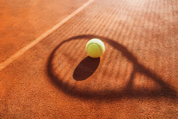 A tennis ball resting on a sunlit clay court casting a shadow from a racquet