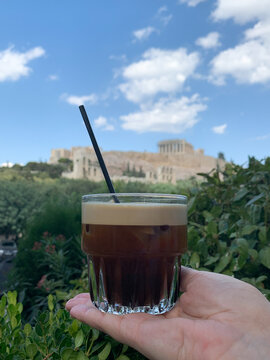 Hand holding a glass of freddo espresso with a black straw against the backdrop of the Acropolis in Athens.