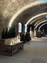 Historic wine cellar with arched brick ceilings, wooden crates, and large glass bottles. Dim...