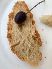 Crispy slice of bread with a dark purple olive on a twig resting on a white plate. Rustic texture, airy crumbs, and golden crust.