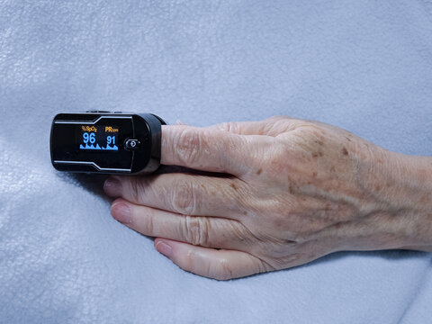 Senior woman patient wearing fingertip pulse oximeter device during medical examination, resting hand on hospital bed, showing blood oxygen saturation level and pulse rate measurement