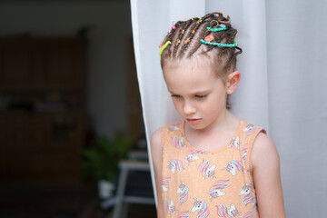 A small cute smiling girl with colorful dreadlock braids standing near a white wall. A concept of a...
