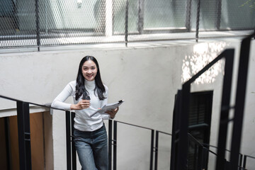 Young professional woman standing confidently on stairs with coffee and documents, smiling in a modern office.