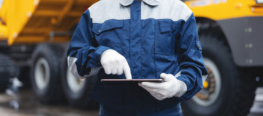 Big yellow dump truck driver man in uniform with tablet computer controls loading of cargo or coal. Concept banner automated logistics online internet
