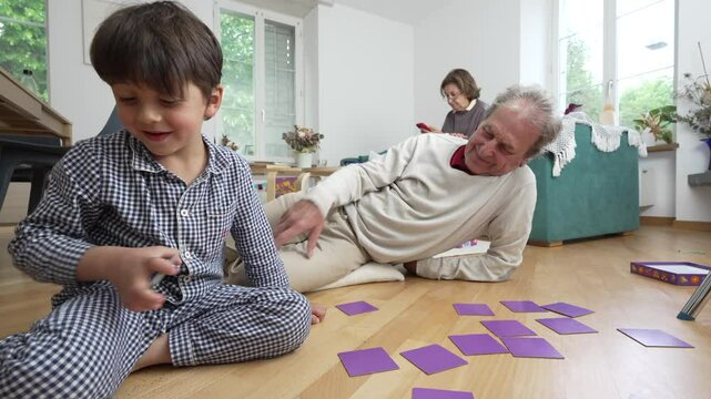 Young boy and grandfather playing memory game on floor, smiling and bonding, grandmother in background with smartphone, happy family time, intergenerational connection, cozy indoor activity