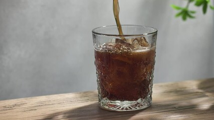 fresh sparkling cola drink pouring into a glass on sunny wooden kitchen tabletop and gray wall background, slow motion