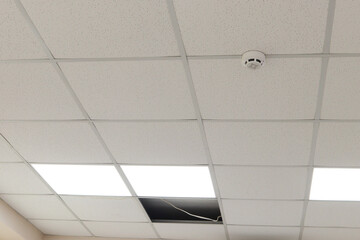 ceiling with a suspended system made of white acoustic panels in a metal frame. such ceilings in office . there is a fire alarm. you can see the removed panel through which the cable passes. 