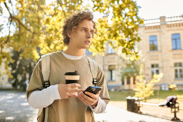 Fototapeta premium Stylish young man enjoys a sunny day while holding coffee and checking his phone in the park