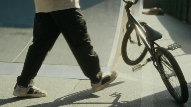 Low section shot of BMX rider picking up bike from ground and riding away while training in sunlit skate park