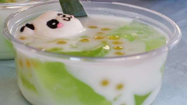 Asian dessert with coconut milk, pandan jelly in plastic cup. A cute panda-shaped mochi or marshmallow floats on top, with golden tapioca pearls. Traditional tropical chinese sweet snack