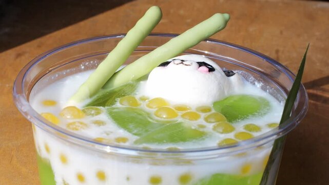 Asian dessert with coconut milk, pandan jelly in plastic cup. A cute panda-shaped mochi or marshmallow floats on top, with golden tapioca pearls. Traditional tropical chinese sweet snack