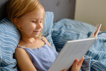 Inquisitive child kneels on rumpled bedsheets analyzing picture book, blue blanket cape-style over...