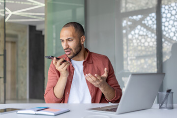 Worried businessman using voice assistant on his smartphone while sitting at his desk in a modern...