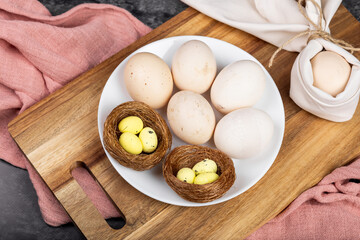 Natural farm fresh eggs on a plate on a dark background, next to decorative painted quail eggs in a nest, flowers, preparation for Easter, painting eggs for Easter, farm concept