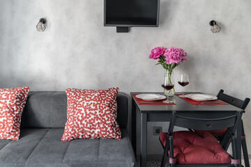 Modern kitchen design. plates, glasses of red wine, and vase of pink peonies on dining table. chairs near table, gray sofa with red cushions, TV. inside home. Modern flat. Floral home decoration.