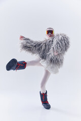 Young woman in dynamic pose in oversized shaggy fur coat, ski goggles, and moon boots against white...