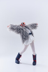 Young woman extending arms in oversized shaggy fur coat, ski goggles, and moon boots against white...