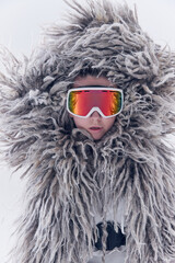 Close-up portrait of young woman in oversized shaggy fur coat and ski goggles against white studio...