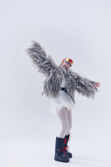 Young woman posing in oversized shaggy fur coat, ski goggles, and moon boots against white studio...