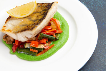 Cod steak on a bed of vegetables in a plate on a dark background.