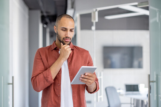 Serious thinking programmer developer standing with tablet computer in hands. Man testing new application software. Confident company employee reading online data.
