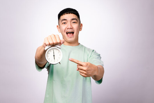 A smiling Asian man in a mint green shirt holds a white alarm clock and points at it with a cheerful expression against a white background, symbolizing time management, punctuality