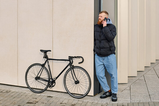 A red-haired man with a beard speaks on the phone and stands near a bicycle