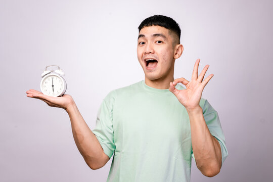 A cheerful Asian man in a mint green shirt holds a white alarm clock in one hand and makes an "OK" gesture with the other, smiling confidently against a white background