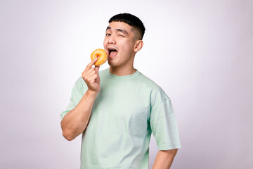 An Asian man in a mint green shirt playfully licks a glazed donut, expressing joy and anticipation....