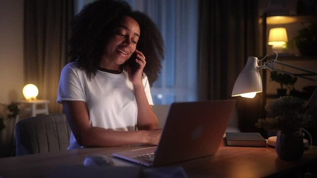A young woman gets a phone call while working on her laptop and smiles happily. She looks engaged and cheerful, enjoying the conversation in a cozy home office at night. Nighttime setting