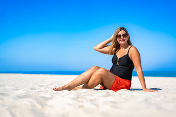 Summer holidays. Portrait of middle aged beautiful woman sitting on white sandy beach on sunny day