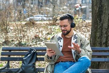 Obraz premium Young hipster businessman with cool attitude sitting in the park having video call on digital tablet. Expatriate business person standing out of the crowd, intelligent design professional occupation.