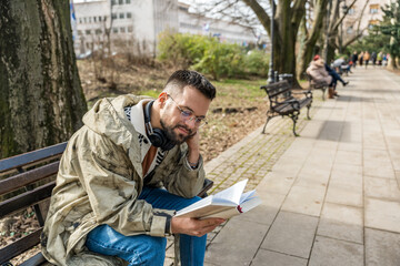 Young curious religious man, reading a book about religion, while sitting in the park on bench. Finding God male, happy and hungry for knowledge about faith. Christian or muslim read Bible or Quran.