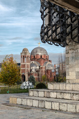 A partial view of the incomplete Christ the Saviour Cathedral in Pristina, Kosovo, framed by a modern architectural element. The cathedral stands against a partially cloudy sky