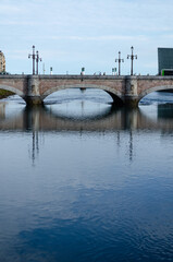 A serene view of the Santa Catalina Bridge in San Sebastián, Spain. The bridge's elegant arches are reflected in the calm waters of the Urumea River, creating a sense of peace and tranquility