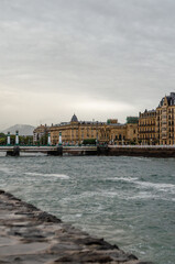 A serene view of San Sebastián and the Urumea River. The image captures the elegant architecture against a cloudy sky, with the river's gentle flow in the foreground