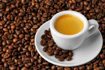 Small white espresso cup overflowing with rich coffee, resting on scattered roasted beans, evoking warm morning brewing ambiance