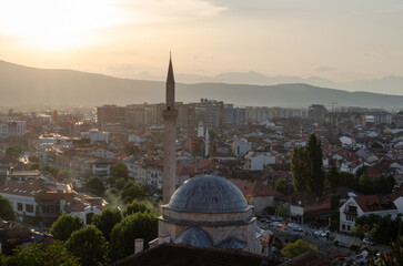 A mosque with a prominent minaret and dome overlooks the cityscape of Prizren, Kosovo, at sunset. The warm light of the setting sun bathes the city in a golden glow