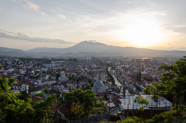 Panoramic view of Prizren, Kosovo, at sunset. The cityscape is nestled between lush green hills, with a historic mosque dome visible amidst the rooftops. A hazy mountain looms in the distance