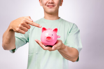 A cheerful Asian man in a mint green shirt smiles while holding and pointing at a pink piggy bank...
