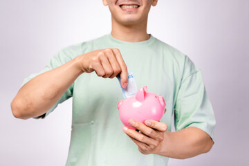 A cheerful Asian man in a mint green shirt smiles while holding a pink piggy bank and inserting a...
